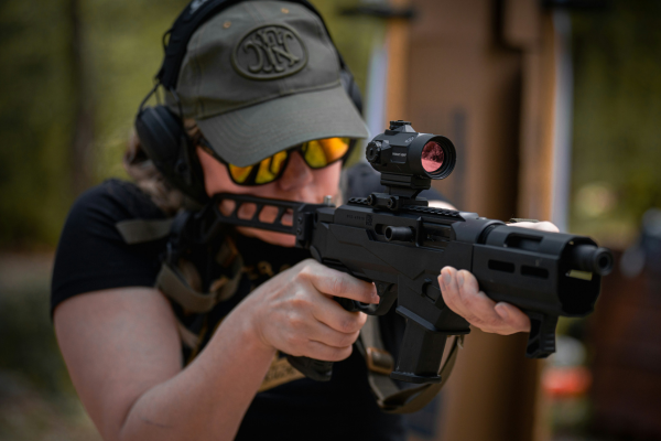 Person aiming a tactical rifle equipped with a red dot optic while wearing eye and hearing protection during outdoor firearms training.